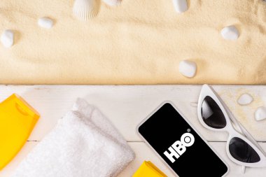 KYIV, UKRAINE - MARCH 25, 2020: Top view of smartphone with hbo app near sunglasses, sunscreen and towel on white wooden planks near pebbles on sand  clipart