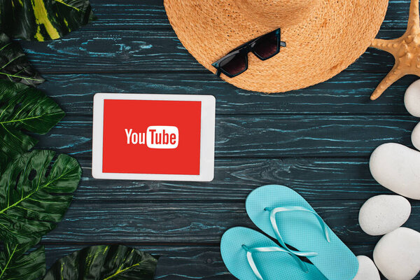 КИЕВ, УКРАИНА - 25 марта 2020 года: Верхний вид цифрового планшета с приложением youtube рядом с шлепанцами, солнцезащитные очки с листьями и морскими камнями на темном деревянном фоне
