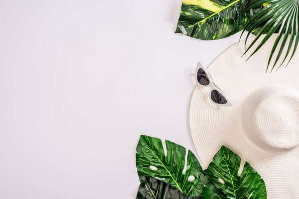 Top view of sunglasses and sun hat near green leaves of tropical plants on white background