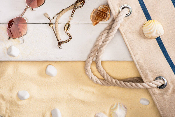 Top view of sunglasses, bracelet and bag on white wooden planks near pebbles on sand