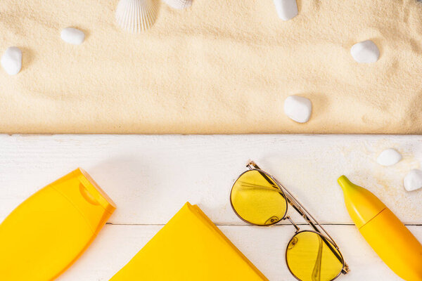 Вид сверху на желтые солнцезащитные кремы, книгу и солнцезащитные очки на белых деревянных досках рядом с морскими камнями и морем на песке
