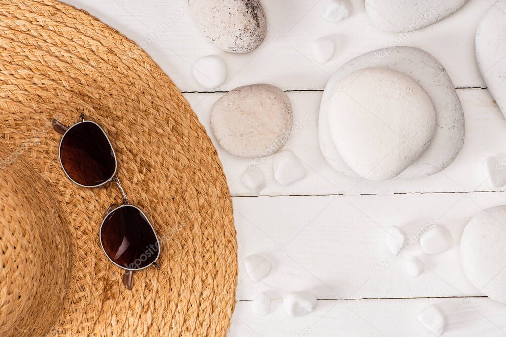 Top view of sunglasses on straw hat near sea stones on white wooden background