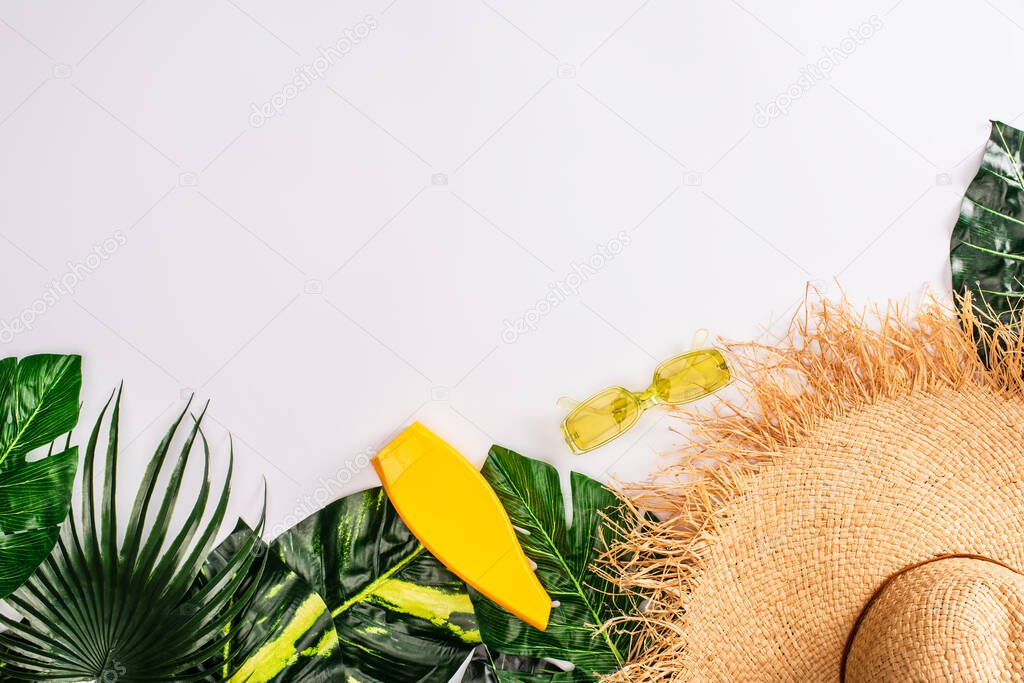 Top view of bottle of sunscreen near sunglasses and straw hat on green leaves on white surface