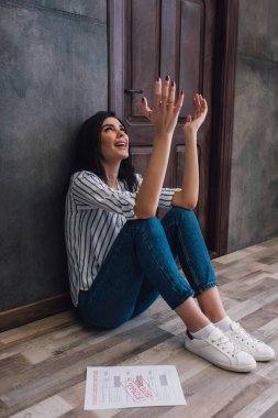 Stressed woman with raised hands looking up on floor near wall in room clipart