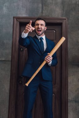 Elinde beysbol sopası olan öfkeli bir koleksiyoncu, parmağıyla işaret ediyor ve odanın kapısında bağırıyor.