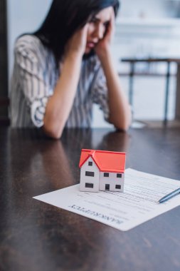 Selective focus of upset woman at table with house model and pen on paper with bankruptcy inscription clipart