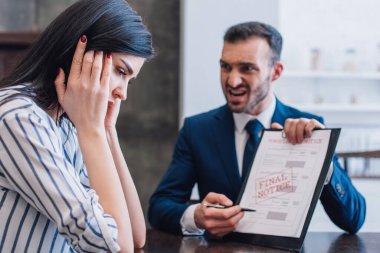 Selective focus of stressed woman and angry collector pointing with pen at documents with final notice lettering at table clipart