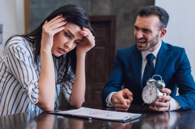 Angry collector holding alarm clock and pointing at documents near stressed woman at table in room clipart