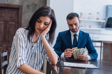 Selective focus of upset woman near documents with collector counting dollar banknotes at table in room clipart