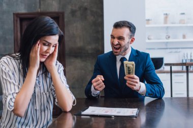Excited collector with dollar banknotes near stressed woman at table in room clipart