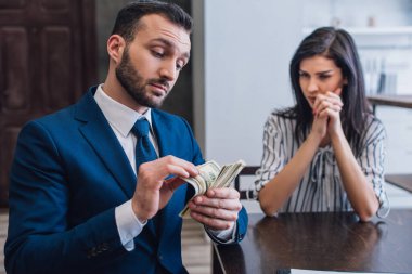 Woman with clenched hands looking at collector counting money at table in room clipart