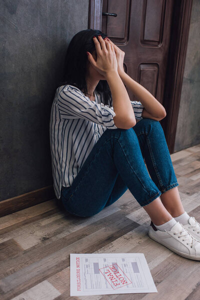 Женщина покрывает лицо рядом с документом с лишением права выкупа и последнее уведомление надпись на полу рядом со стеной в комнате
