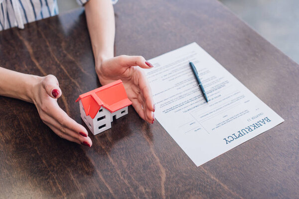Обрезанный вид женских рук рядом с моделью дома, бумага с надписью о банкротстве и ручка на столе
