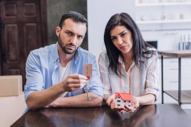 Adam üzgün bir kadının yanında kameraya bakıyor ve anahtarı satılık mektuplarla ve masadaki ev modeliyle gösteriyor.