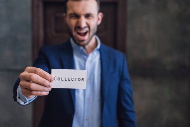 Selective focus of angry collector shouting and showing card with collector lettering in room clipart
