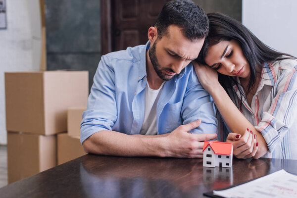 Woman leaning to upset husband at table with house model and documents in room