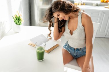 Çekici kız, mutfakta bir bardak smoothie içerken kitap okurken gülümsüyor. 