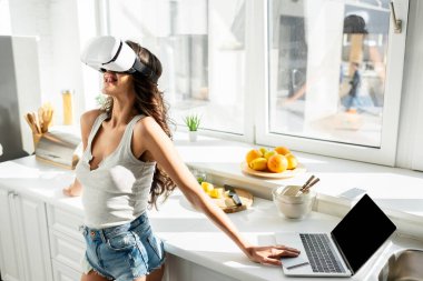 Young woman in denim shorts sticking out tongue while using vr headset near laptop and credit card in kitchen 