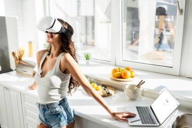 KYIV, UKRAINE - MARCH 13, 2020: Young woman using vr headset near laptop with google website and credit card in kitchen  clipart
