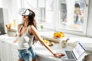 KYIV, UKRAINE - MARCH 13, 2020: Woman in vr headset standing near laptop with facebook website and credit card on kitchen worktop  clipart