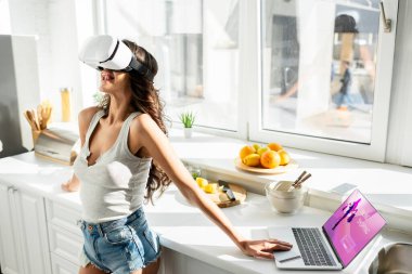 İnternette alışveriş sitesi ve mutfakta kredi kartı olan, dizüstü bilgisayarın yanında vr kulaklık kullanan kot şortlu bir kız. 