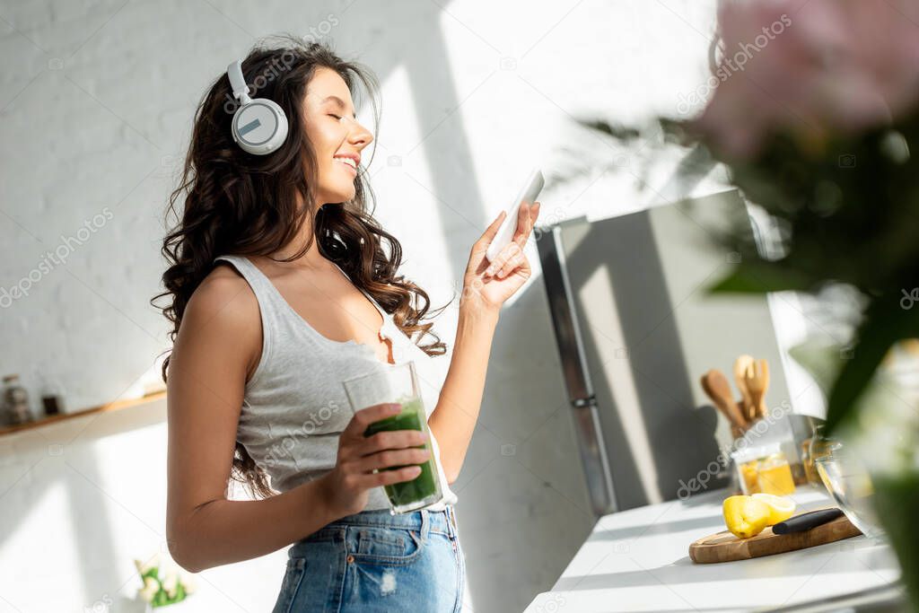 Selective focus of smiling woman in headphones holding smartphone and glass of smoothie in kitchen 
