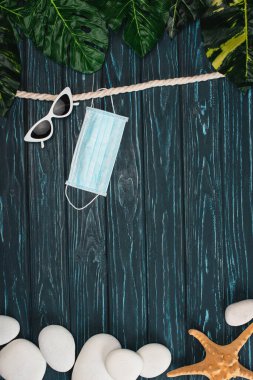 Tıbbi maskenin, güneş gözlüklerinin ve çakıl taşlarının ve deniz yıldızlarının yakınındaki kara tahta yüzeyin üst görüntüsü.