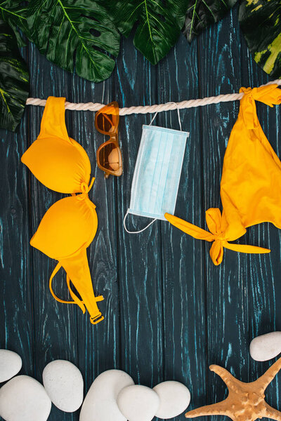 Top view of yellow swimsuit near medical mask, sunglasses and leaves on dark wooden surface