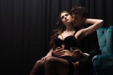 İç çamaşırlı seksi bir kadın elinde bir bardak viskiyle yakışıklı bir adamın üzerine oturuyor.