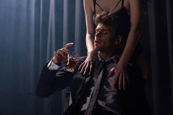 seductive woman touching man sitting and holding glass of whiskey on black