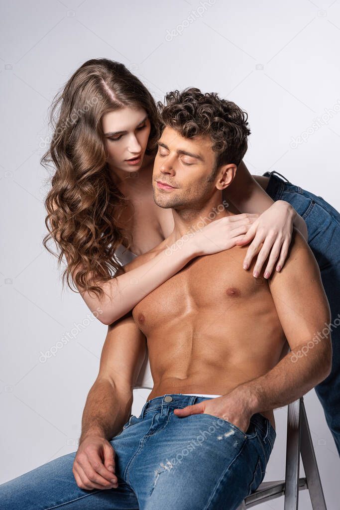 attractive young woman touching shirtless man in jeans sitting with hand in pocket on grey 