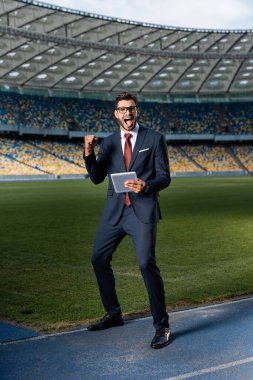 young businessman in suit and glasses using digital tablet and showing yeas gesture at stadium, sports betting concept clipart