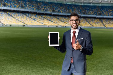 Takım elbiseli, gözlüklü, elinde boş ekranlı dijital tablet ve stadyumda baş parmağını gösteren mutlu genç iş adamı.