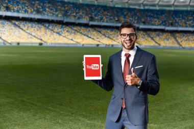 KYIV, UKRAINE - 20 Haziran 2019: Takım elbiseli mutlu genç işadamı ve elinde dijital tablet ve youtube uygulaması ile stadyumda baş parmağını gösteriyor