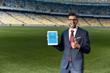 KYIV, UKRAINE - 20 Haziran 2019: Takım elbiseli mutlu genç işadamı ve Skype uygulamalı dijital tablet tutan ve stadyumda baş parmağını gösteren gözlüklü genç adam