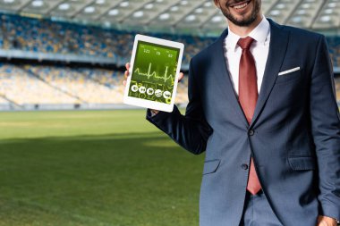 Takım elbiseli gülümseyen genç işadamının, stadyumdaki sağlık uygulaması ile dijital tablet tutarken çekilmiş görüntüsü.