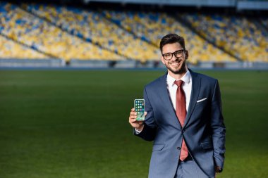 KYIV, UKRAINE - 20 Haziran 2019: Takım elbiseli gülümseyen genç işadamı ve stadyumdaki uygulamalı akıllı telefon gözlüğü