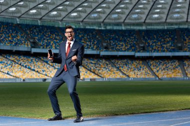 full length view of excited young businessman in suit and glasses holding smartphone with blank screen and showing yes gesture at stadium clipart