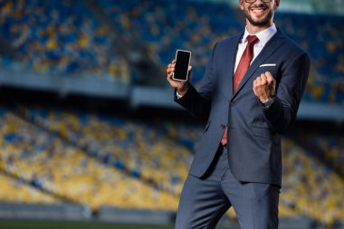 cropped view of smiling young businessman in suit holding smartphone with blank screen and showing yes gesture at stadium clipart