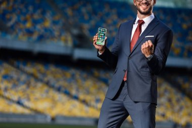 KYIV, UKRAINE - 20 Haziran 2019: takım elbiseli gülümseyen genç işadamının iPhone uygulamalarıyla akıllı telefon tutuşu ve stadyumda evet hareketi göstermesi