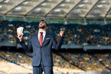 young businessman in suit and glasses with money showing yeas gesture and yelling at stadium clipart
