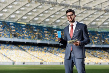 KYIV, UKRAINE - 20 Haziran 2019: Stadyumdaki Skype uygulamalı akıllı telefonu gösteren takım elbiseli ve gözlüklü gülümseyen genç işadamı