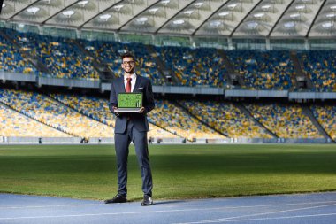 Takım elbiseli gülümseyen genç işadamı stadyumdaki futbol formasyonunda dizüstü bilgisayarı gösteriyor.