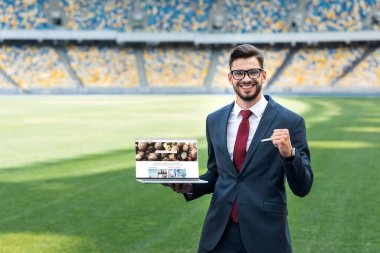 KYIV, UKRAINE - 20 Haziran 2019: Stadyumdaki depozito fotoğrafları web sitesinde dizüstü bilgisayarını tutarken takım elbiseli gülümseyen genç işadamı evet işareti yapıyor