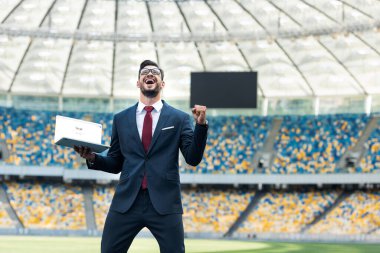 KYIV, UKRAINE - 20 Haziran 2019: Takım elbiseli mutlu genç işadamının düşük açılı görüntüsü evet hareketi gösteriyor ve google web sitesi ile stadyumda dizüstü bilgisayarı tutuyor