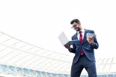 low angle view of happy young businessman in suit holding laptop and money at stadium, sports betting concept clipart