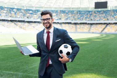 Futbol sahasında oturmuş futbol topuyla takım elbiseli gülümseyen genç işadamı, spor bahisleri konsepti.