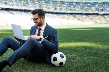 Futbol sahasında oturmuş futbol topuyla takım elbiseli gülümseyen genç işadamı, spor bahisleri konsepti.