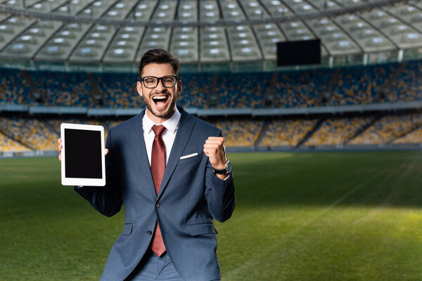 cheerful young businessman in suit and glasses holding digital tablet with blank screen and showing yes gesture at stadium