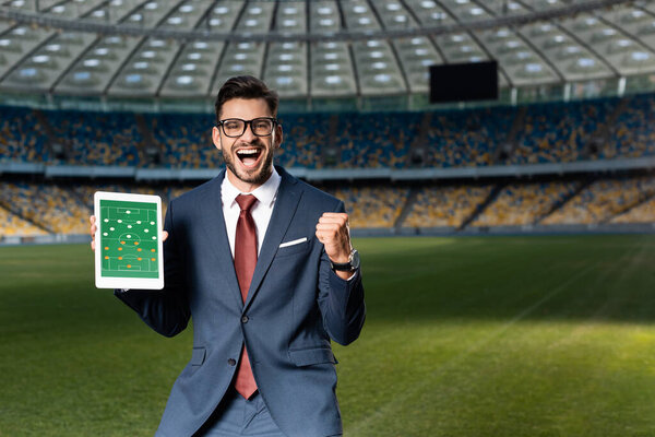 cheerful young businessman in suit and glasses holding digital tablet with soccer game on screen and showing yes gesture at stadium